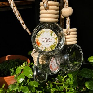 Könnte beinhalten: Eine Gruppe runder, transparenter Glasflaschen für Auto-Lufterfrischer mit hellen Holzverschlüssen und geflochtenen Aufhängeschnüren. Jede Flasche hat ein rundes Etikett mit der Aufschrift Mango & Gardenia, verziert mit Blumen- und Blattmotiven.