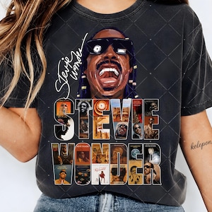 May include: A charcoal gray t-shirt featuring a large graphic of Stevie Wonder wearing sunglasses with his mouth open. The design includes the text Stevie Wonder in script and a collage of various album covers arranged behind the name.
