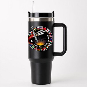 May include: A matte black stainless steel tumbler with a handle, a clear straw, and a circular graphic featuring a spacecraft, a sunburst, and international flags surrounding the text PROJECT HAIL MARY in white capital letters.