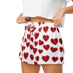 May include: White shorts featuring a repeating pattern of solid red hearts. The shorts have an elastic waistband with a matching white drawstring tie at the center front. The fabric has a smooth, slightly reflective finish throughout the garment.