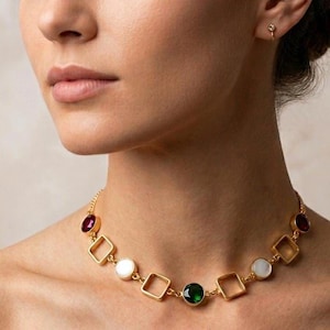 Puede incluir: Un collar de cadena en tono dorado con elementos geométricos alternos, incluyendo marcos cuadrados abiertos y piedras circulares engastadas en rojo intenso, verde esmeralda y blanco iridiscente, junto a un pendiente a juego.