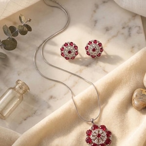 Könnte beinhalten: Ein Schmuckset bestehend aus einer silberfarbenen Kettenhalskette mit rundem Anhänger und passenden runden Ohrsteckern. Jedes Stück zeigt eine zentrale Anordnung klarer Steine, umgeben von einem Ring aus leuchtend roten, rund geschliffenen Steinen.
