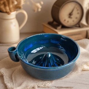 Könnte beinhalten: Eine tiefblaue Zitruspresse aus Keramik mit einem zentralen Presskegel und einem kleinen seitlichen Griff. Die Schale hat eine glänzende, nuancierte Oberfläche in verschiedenen Blautönen und dient zum Auspressen von Zitronen oder Orangen.