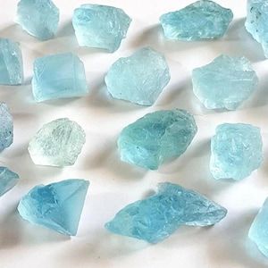 Peut inclure: Une collection de dix-neuf pierres d'aigue-marine brutes et bleu clair dispersées sur une surface blanche unie. Chaque pierre présente une forme irrégulière et dentelée avec des propriétés translucides et des tailles variées.