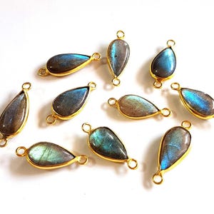 May include: A collection of ten teardrop-shaped labradorite gemstones, each encased in a gold-toned metal bezel setting. Every piece features two small circular loops at opposite ends, displaying iridescent blue and gray flashes of color.