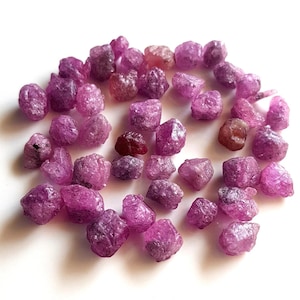 Op de afbeelding: Een verzameling kleine, ruwe, ongepolijste robijn-edelstenen in verschillende tinten dieproze en magenta. Elke steen heeft een ruw, onregelmatig en getextureerd oppervlak met natuurlijke facetten en kristallijne structuren verspreid over het beeld.