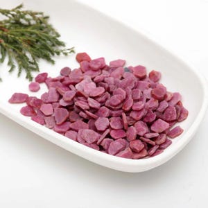 Puede incluir: Una colección de pequeñas astillas de rubí planas e irregulares en tonos rosa intenso y magenta. Las piedras están amontonadas en un plato blanco, alargado y curvo, junto a una pequeña rama de follaje de cedro verde.