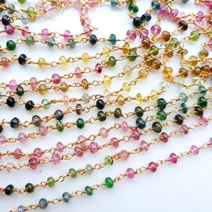 Op de afbeelding: Een meerstrengs gouden ketting met kleine, gefacetteerde, veelkleurige toermalijnkralen in tinten roze, groen, geel en zwart. De kralen zijn individueel omwikkeld met draad en verbonden door dunne gouden metalen schakels in een verfijnd ontwerp.