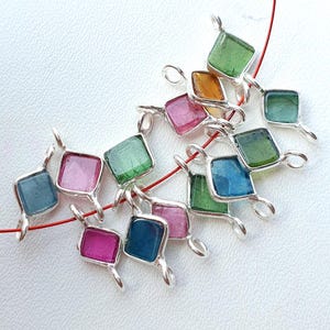 May include: A collection of small, square-shaped jewelry charms featuring translucent, multi-colored stones set in silver-toned metal frames. Each charm has two small loops on opposite sides, allowing them to be strung together on a thin red cord.