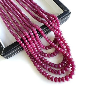 May include: A multi-strand necklace featuring small, round, deep magenta-colored beads. The beads have a smooth, polished finish and are strung together in several cascading layers, creating a dense, textured appearance of vibrant pinkish-red stones.