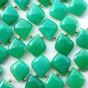 Könnte beinhalten: Eine Ansammlung von facettierten, rautenförmigen grünen Edelsteinperlen, die in Reihen angeordnet und mit kleinen, runden, metallisch goldfarbenen Zwischenperlen zwischen jedem einzelnen grünen Stein verbunden sind.