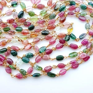 May include: A multi-strand necklace featuring small, oval-shaped, polished tourmaline gemstones in various shades of pink, green, and yellow. Each gemstone is individually linked together with thin, gold-toned wire, creating a delicate chain.