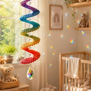 Op de afbeelding: Een hangende gehaakte mobiel met een spiraalvormige structuur in regenboogkleuren, bekroond met een witte pluche wolk en een oranje zon. Heldere gefacetteerde kristalkralen zijn in het midden geregen en eindigen in een groot kristallen prisma.