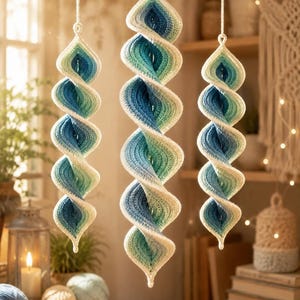 Op de afbeelding: Drie hangende spiraalvormige ornamenten gemaakt van gehaakt garen. Elk stuk heeft een kleurverloop van diepblauw in het midden naar lichtblauw, turquoise en wit aan de buitenranden, opgehangen aan lussen van wit garen.