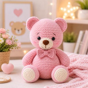 Può includere: Un orsacchiotto all'uncinetto rosa con un papillon rosa abbinato, un muso color crema, un naso ricamato marrone e toppe circolari color crema sulle piante dei piedi. L'orso ha occhi di sicurezza neri e sopracciglia ricamate.