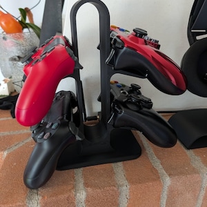 May include: A black plastic vertical gaming controller stand holding four controllers. On the left side are one red and one black controller. On the right side are one metallic red and one black controller, each featuring multi-colored buttons.