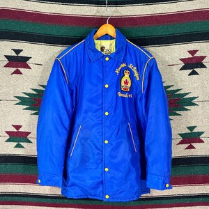 May include: A bright blue coach jacket featuring a yellow snap-button front closure, a pointed collar, and long sleeves. The chest displays an embroidered emblem with a crown and maple leaf, accompanied by the text Norwood St. Boniface Branch #43.