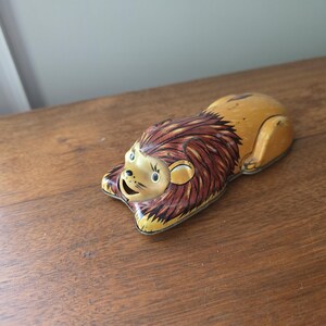 Pode incluir: Um brinquedo de lata vintage em forma de leão na posição agachada, com corpo amarelo e juba castanha. O leão apresenta detalhes faciais pintados, incluindo olhos pretos, nariz e boca aberta. A superfície metálica mostra sinais de desgaste e pátina.
