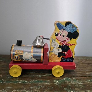 Puede incluir: Tren de juguete de madera vintage con Mickey Mouse. El tren tiene una base roja, ruedas amarillas y un motor plateado con el texto "No. 465 Choo-Choo". Mickey Mouse está representado en un recorte de madera, con gorro azul y bufanda roja.