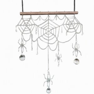 May include: A decorative wall hanging featuring a horizontal wooden rod with a black chain for suspension. Three intricate spider web patterns made of clear, shimmering beads are suspended from the rod, accented with three beaded spiders and clear crystal spheres.