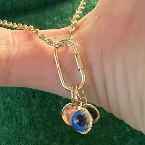 Op de afbeelding: Een goudkleurige ketting met een gedraaide schakelketting en een langwerpige, ovale karabijnsluiting. Aan de sluiting hangen twee kleine ronde bedels, waarvan er één een blauw en zwart oogontwerp heeft met een textuurrijke metalen rand.
