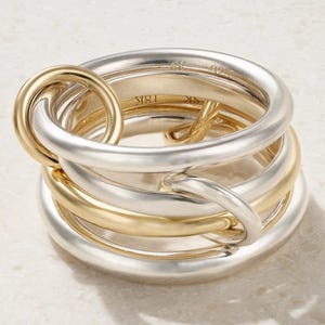 May include: A multi-band ring featuring interlocking silver and gold-toned circular bands. The interior of the gold band is clearly engraved with the text 18K, while the silver band features an engraved hallmark. The design consists of several smooth, rounded metal loops that overlap to create a layered, cohesive structure.