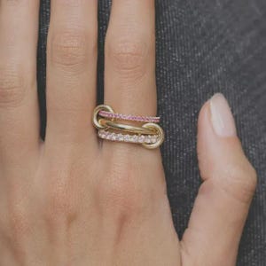 Pode incluir: Um anel de ouro com três bandas entrelaçadas. Uma banda é cravejada com pequenas pedras cor-de-rosa, outra banda com pequenas pedras transparentes e a terceira banda é uma banda de metal dourado polido. Os anéis formam um elo contínuo.