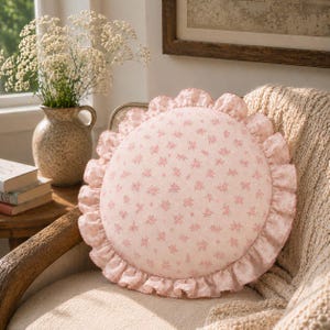 May include: A round throw pillow featuring a light pink fabric with a small-scale floral pattern in a darker pink hue. The pillow is finished with a gathered, ruffled edge made from the same floral fabric, creating a soft, circular silhouette.
