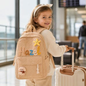 Może przedstawiać: Beżowy plecak sztruksowy z wyhaftowanym imieniem Hazel w pomarańczowe i niebieskie litery. Torba ozdobiona jest żółtą pluszową naszywką w kształcie gwiazdki na górze oraz małą pluszową naszywką z pieskiem na przedniej kieszeni z białym chwostem.