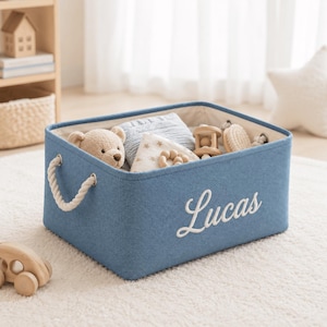 Puede incluir: Una cesta de almacenamiento rectangular de tela azul con asas de cuerda blanca y el nombre Lucas bordado en letra cursiva blanca en la parte frontal. La cesta contiene varios artículos de bebé, incluyendo un oso de peluche, juguetes de madera y textiles.
