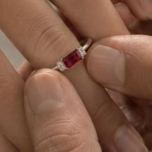 Puede incluir: Un anillo de tono plateado que presenta una piedra preciosa roja de corte rectangular tipo baguette en el centro, flanqueada por dos pequeñas piedras de acento transparentes de corte redondo a cada lado, sobre una banda de metal pulido.
