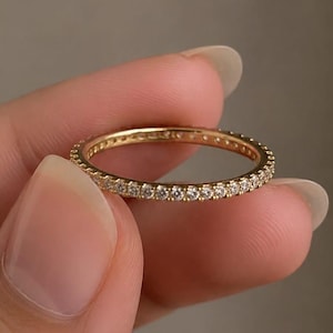 Peut inclure: Une fine bague en or dotée d'une rangée continue de petites pierres claires taillées en rond, serties sur toute la circonférence extérieure. Le métal présente une finition polie et réfléchissante, avec des pierres régulièrement espacées.