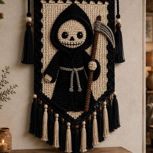 May include: A crocheted wall hanging featuring a central figure of a hooded skeleton holding a scythe. The design uses black and cream yarn, accented with wooden beads and black and cream tassels hanging from a wooden rod with a black cord.