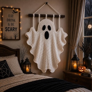 May include: A white crocheted ghost wall hanging suspended from a dark wooden branch with a rope. The ghost features two black oval eyes and a larger black oval mouth, creating a classic spectral shape with wavy, textured edges for display.