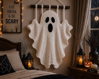 May include: A white crocheted ghost wall hanging suspended from a dark wooden branch by four loops. The ghost features two black oval eyes and a larger black oval mouth, creating a classic spectral shape with wavy, textured edges throughout.