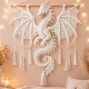 Puede incluir: Un gran tapiz de macramé en color blanco roto que presenta un intrincado diseño de dragón con alas extendidas, una larga cola enroscada y nudos detallados. La pieza cuelga de una varilla de madera horizontal y está decorada con borlas y cuentas.