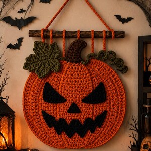 Könnte beinhalten: Ein runder, gehäkelter Halloween-Kürbis als Wandbehang in Orange mit einem dunkelbraunen Stiel, einem grünen Blatt und einer grünen Ranke. Das Gesicht besteht aus schwarzen gehäkelten Augen, einer Nase und einem gezackten Mund.