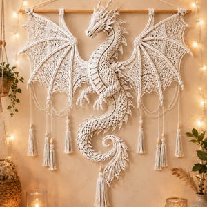 Peut inclure: Une grande tenture murale en macramé blanc cassé représentant un dragon détaillé avec des ailes déployées, une longue queue courbée et des motifs noués complexes. Le dragon est suspendu à une tige en bois et orné de plusieurs pompons.