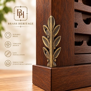 May include: A dark brown wooden cabinet corner featuring an attached antique brass leaf-shaped decorative accent. The brass piece has a vertical stem with symmetrical leaves and two visible screw holes. Text on the image reads BRASS HERITAGE.