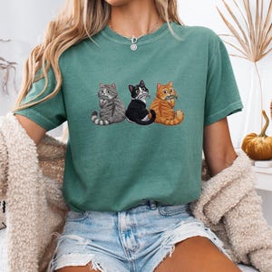 Può includere: Una t-shirt a maniche corte color verde salvia con una stampa grafica di tre gatti seduti in fila. I gatti sono raffigurati con motivi tigrato grigio, bianco e nero, e tigrato arancione. La t-shirt presenta un girocollo.