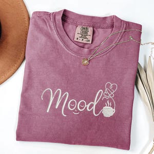 May include: A mauve short-sleeve t-shirt featuring the word "mood" embroidered in white thread, with the letter "o" stylized as a smiling face and a steaming coffee cup graphic with a heart-shaped steam trail. The shirt has a Comfort Colors label.