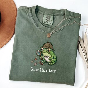 Puede incluir: Una camiseta verde oliva con un bordado de una rana verde que lleva un sombrero de detective y gafas, sosteniendo una lupa y un bloc de notas. El texto "Bug Hunter" está bordado debajo de la rana, junto con pequeños insectos.