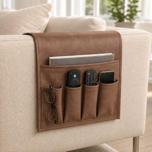 Op de afbeelding: Een bruine kunstlederen armleuning-organizer met een groot bovenvak met een notitieboek en vier kleinere voorvakken met een bril, twee afstandsbedieningen en een smartphone, hangend over de armleuning van een beige bank.