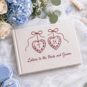 Puede incluir: Un libro de visitas rectangular de color blanco roto con detalles bordados en granate. La portada presenta dos adornos en forma de corazón con las letras T y H en su interior, atados con lazos. El texto Letters to the Bride and Groom aparece.