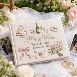 Puede incluir: Un libro de visitas de boda de tela color crema con bordados de un pastel de tres niveles, una botella de champán, dos copas, anillos de boda, un lazo, un sobre con sello de cera y un ramo de flores, con el texto Mr & Mrs, Noah & Olivia, September 21, 2027.