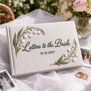 Puede incluir: Un libro rectangular cubierto de tela color hueso con hojas verdes y flores de lirio de los valles blancas bordadas. El texto "Letters to the Bride" está bordado en cursiva verde oscuro, seguido de la fecha "10.12.2027" en verde oscuro.