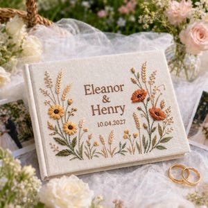 Puede incluir: Un álbum de fotos rectangular cubierto de tela color hueso con motivos florales bordados en tonos amarillo, naranja y verde. El centro muestra el texto bordado Eleanor & Henry y la fecha 10.04.2027 en hilo de color marrón.