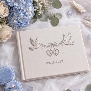 Puede incluir: Un libro de visitas rectangular cubierto de tela color crema con un diseño bordado de dos palomas que sostienen una cinta conectada a dos corazones con las letras S y L, y la fecha 09.18.2027 bordada en hilo oscuro en la portada.
