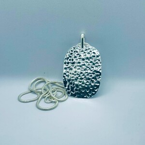 May include: A silver-colored pendant with a textured, hammered surface and an oval shape, attached to a silver-colored snake chain. The pendant features a reflective, dimpled finish that catches the light, creating a high-contrast appearance.