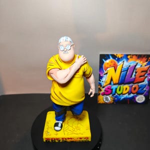 Puede incluir: Una figura coleccionable de un personaje con cabello blanco y gafas redondas, vistiendo una camiseta amarilla brillante y pantalones azules. La figura se apoya en una base cuadrada amarilla con texto, junto a una tarjeta con el texto NILE STUDIOZ.